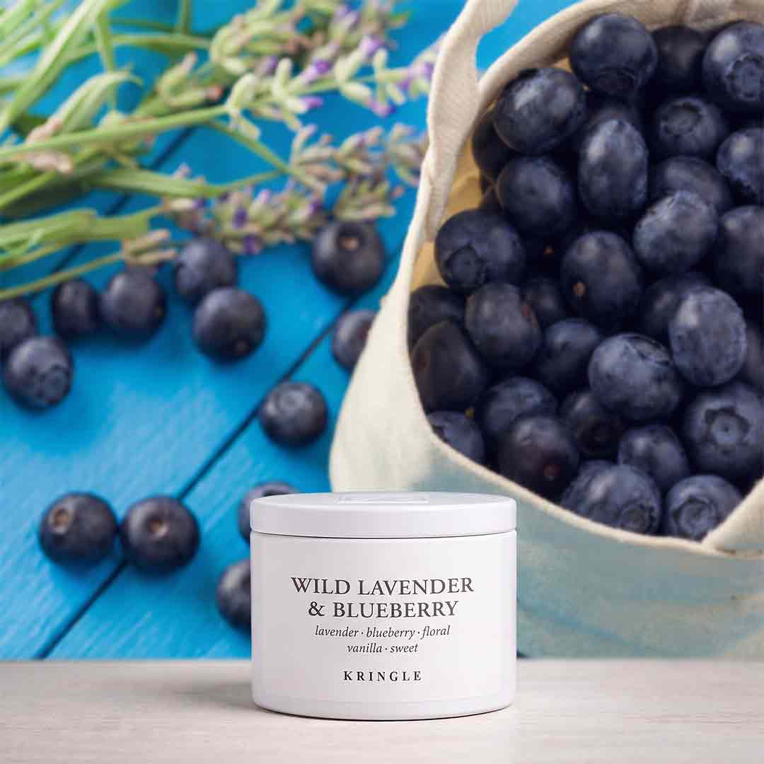 A white jar of KRINGLE Wild Lavender & Blueberry hair product sits on a light wood surface, styled with fresh blueberries in a canvas sack and lavender sprigs on a bright blue wooden background.