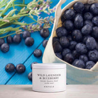 A white jar of KRINGLE Wild Lavender & Blueberry hair product sits on a light wood surface, styled with fresh blueberries in a canvas sack and lavender sprigs on a bright blue wooden background.