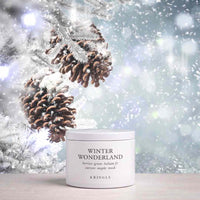 A white cylindrical jar of KRINGLE Winter Wonderland berries green halmus for current maple mask sits on a light wooden surface. The background is a festive, snowy winter scene with pinecones on frosted branches and soft bokeh lights.