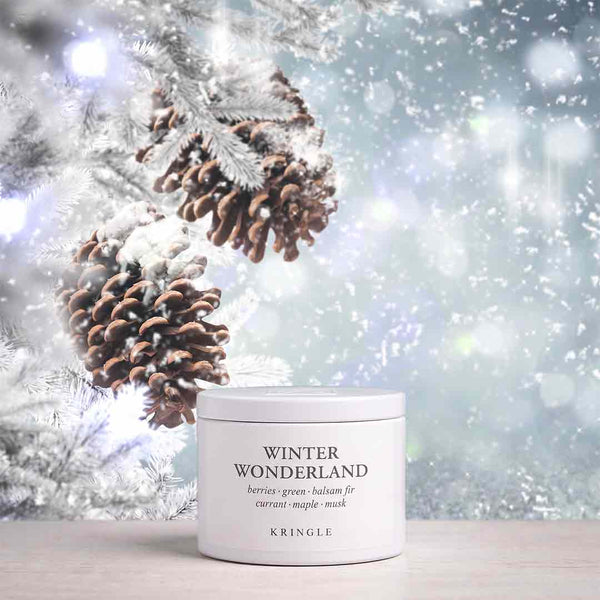 A white cylindrical jar of KRINGLE Winter Wonderland berries green halmus for current maple mask sits on a light wooden surface. The background is a festive, snowy winter scene with pinecones on frosted branches and soft bokeh lights.