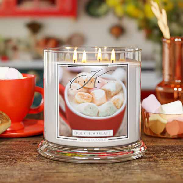 A lit glass jar candle labeled 'Kringle HOT CHOCOLATE' sits on a wooden table in a cozy, blurred kitchen-like setting with a red mug, marshmallows, and warm background decor.