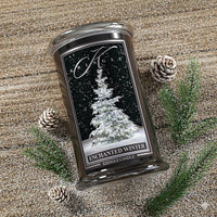 Kringle Candle Enchanted Winter candle on a textured surface with pinecones and branches.