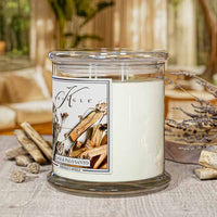 A candle in a glass jar with a label on a table, surrounded by wooden sticks and lavender.