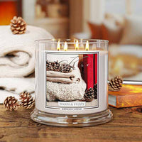 A lit scented candle in a clear glass jar sits on a wooden table, surrounded by cozy props including folded cream knit fabric, pinecones. The front label reads 'WARM & FUZZY' and 'KINGLE CANDLE'.