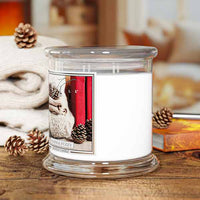 A white scented candle in a clear glass jar with a matching glass lid, styled on a wooden surface with pinecones, a folded cream knit blanket, and a book in a warm indoor setting.