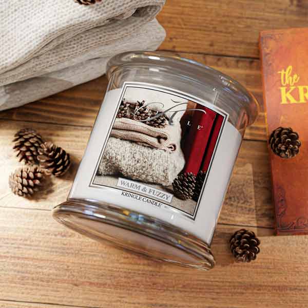 A glass jar candle labeled 'WARM & FUZZY' by KINGLE CANDLE, shown on a wooden surface with folded beige fabric, pinecones, and part of an orange book visible.