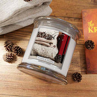 A glass jar candle labeled 'WARM & FUZZY' by KINGLE CANDLE, shown on a wooden surface with folded beige fabric, pinecones, and part of an orange book visible.