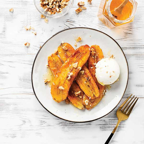Plated dessert with caramelized bananas, nuts, and a scoop of ice cream on a white wooden surface.