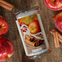 Kringle Candle Spiced Apple candle surrounded by apples and cinnamon sticks on a wooden surface