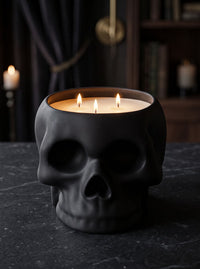 A black skull-shaped candle holder filled with three lit white candles, set on a dark stone surface with a moody, dimly lit background featuring books and candles.