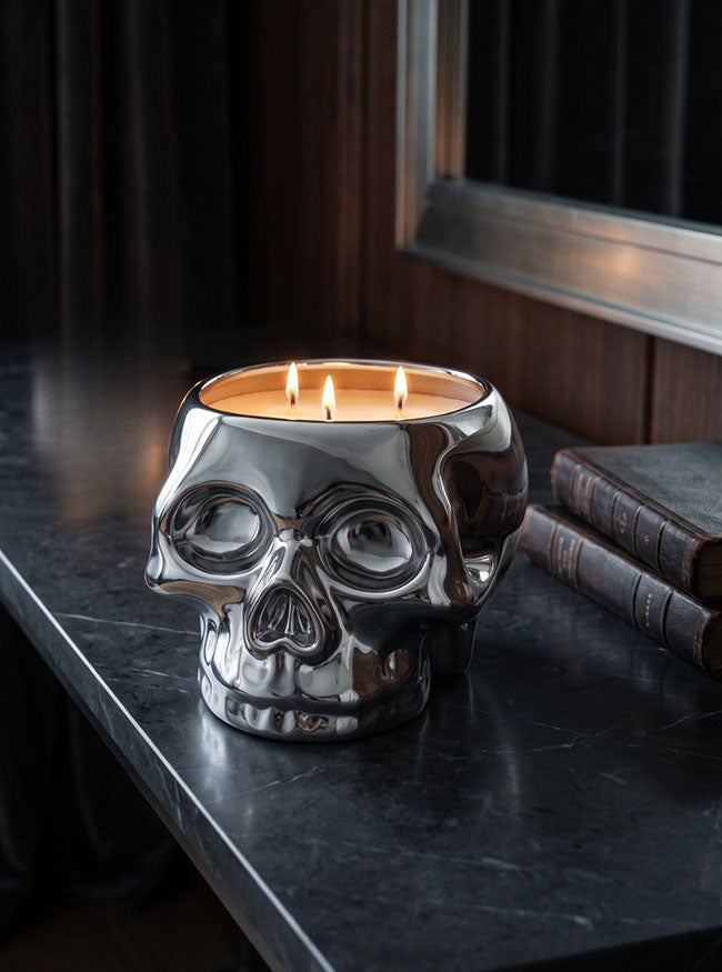 A three-wick candle in a polished silver skull-shaped ceramic holder, lit and set on a dark marble tabletop next to old books.