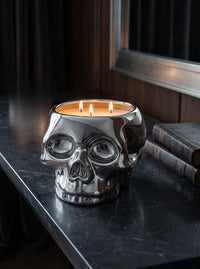 A three-wick candle in a polished silver skull-shaped ceramic holder, lit and set on a dark marble tabletop next to old books.