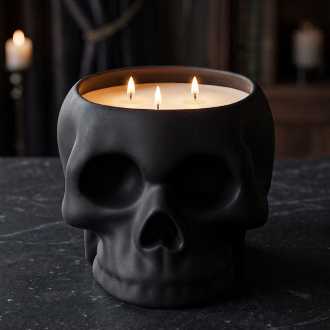 A black skull-shaped candle holder filled with three lit white candles, set on a dark stone surface with a moody, dimly lit background featuring books and candles.