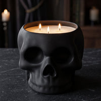 A black skull-shaped candle holder filled with three lit white candles, set on a dark stone surface with a moody, dimly lit background featuring books and candles.