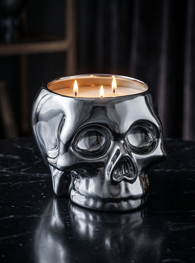 A polished silver skull-shaped candle holder with three lit candles inside, set on a dark reflective surface against a dark draped background.
