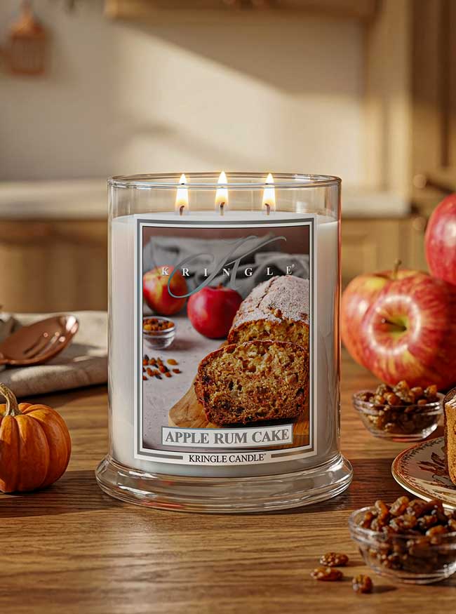 A lit Kringle Candle in a clear glass jar with three flames, labeled Apple Rum Cake, set on a wooden table with apples, nuts, and a small pumpkin in a warm kitchen-like setting.