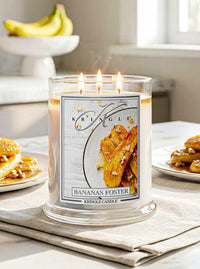 A lit Kringle Candle in a clear glass jar with three flames, labeled 'Bananas Foster,' placed on a table with pancakes and bananas in a softly lit kitchen setting.
