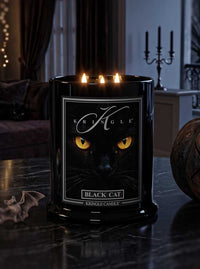 A black glass candle jar with three lit candles, featuring a label of a black cat's face with glowing yellow eyes and the brand name Kringle. The candle is set on a dark marble table in a dimly lit, moody room for Halloween.