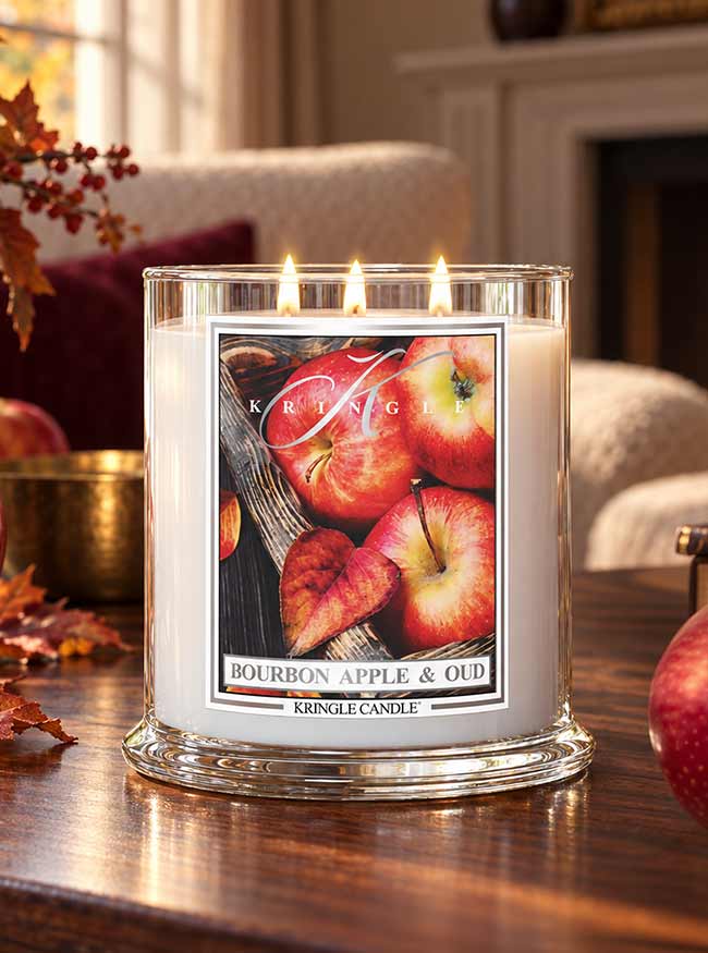 A lit Kringle Candle in a clear glass jar on a wooden table, with a label showing red apples and the scent name Bourbon Apple & Oud. The background shows a cozy living room interior with a fireplace and autumn leaves.