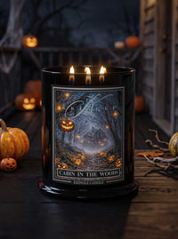 A black Kringle Candle jar with three lit wicks, featuring a Halloween-themed label that reads 'Cabin in the Woods' and shows a glowing haunted cabin scene. The candle is photographed on a dark wooden surface with pumpkins and a spooky outdoor background.