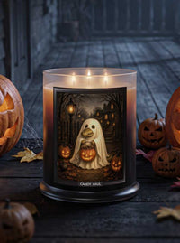 A three-wick candle jar sits on a dark wooden surface surrounded by Halloween decorations, including carved pumpkins and autumn leaves. The candle features a black label with an illustration of a ghostly alligator holding glowing jack-o'-lanterns.