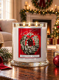 Christmas Stroll 3-wick lit candle on a coffee table in living room with holiday decor and fireplace. Label has a holiday wreath on a door.