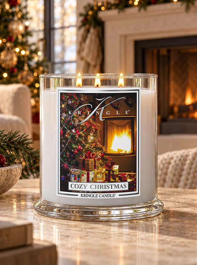 A large glass jar candle labeled 'Cozy Christmas' by Kringle Candle sits on a marble table in a warmly lit living room. The candle has white wax and a festive printed label showing a decorated Christmas tree, wrapped gifts, and a glowing fireplace.