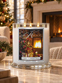 A large glass jar candle labeled 'Cozy Christmas' by Kringle Candle sits on a marble table in a warmly lit living room. The candle has white wax and a festive printed label showing a decorated Christmas tree, wrapped gifts, and a glowing fireplace.