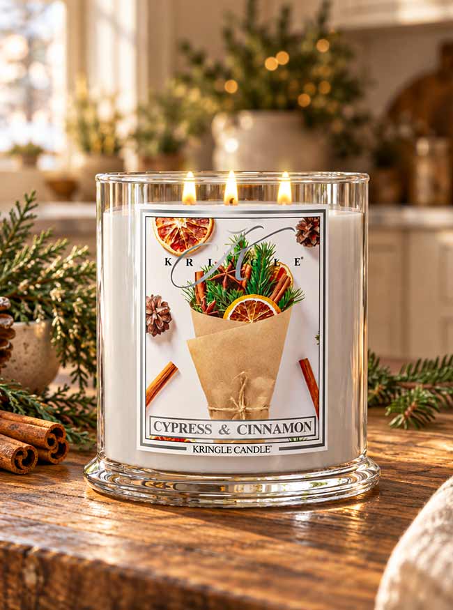 A Kringle Candle jar candle on a wooden table in a cozy indoor setting, with greenery, cinnamon sticks, and warm blurred lights in the background. The candle has a white wax body in a clear glass jar with a festive front label reading 'CYPRESS & CINNAMON' and 'KRINGLE CANDLE.