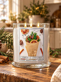 A Kringle Candle jar candle on a wooden table in a cozy indoor setting, with greenery, cinnamon sticks, and warm blurred lights in the background. The candle has a white wax body in a clear glass jar with a festive front label reading 'CYPRESS & CINNAMON' and 'KRINGLE CANDLE.
