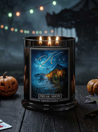 A black glass jar candle with three lit wicks and a Halloween-themed label reading 'Kringle' and 'Freak Show,' placed on a dark wooden surface with pumpkins and blurred lights in the background.