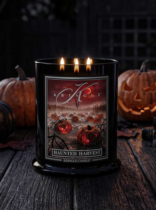 A black Kringle candle labeled 'Haunted Harvest' with three lit wicks, shown on a rustic wooden surface with carved pumpkins and a dark Halloween-themed background.