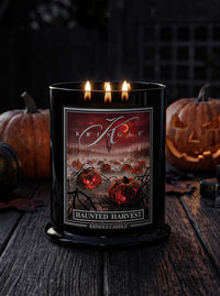 A black Kringle candle labeled 'Haunted Harvest' with three lit wicks, shown on a rustic wooden surface with carved pumpkins and a dark Halloween-themed background.