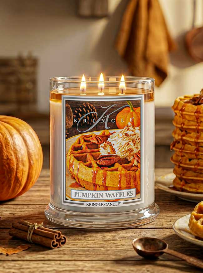 A Kringle Candle in a clear glass jar with three lit wicks, labeled 'Pumpkin Waffles,' set on a rustic wooden table with a pumpkin, cinnamon sticks, and waffle stacks in the background.