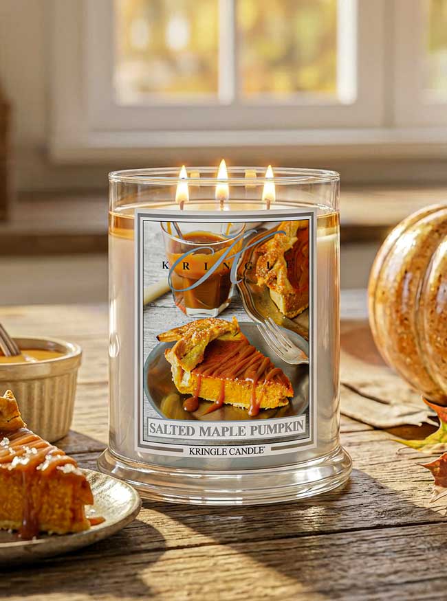 Kringle Candle salted maple pumpkin scented candle with a pie and coffee image on the label. Candle is lit and set on a table with a slice of pie.