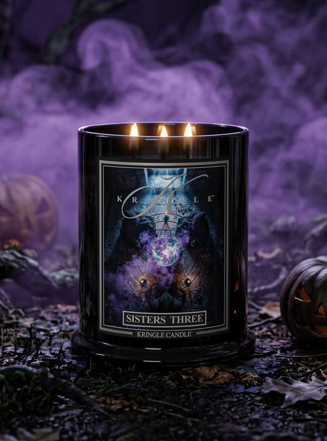 A black glass jar candle with three lit wicks, labeled 'Sisters Three' by Kringle Candle, set in a spooky Halloween scene with purple fog, dried leaves, and jack-o'-lanterns.