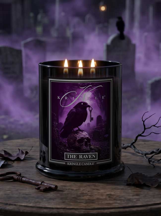 A black glass jar candle with three lit wicks and a front label reading 'The Raven' and 'Kringle Candle,' styled on a wooden surface with dried leaves and branches against a purple misty graveyard background.