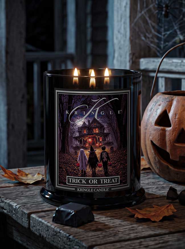A black glass jar candle with three lit wicks sits on a rustic wooden table, surrounded by Halloween props including a carved pumpkin and candy. The front label reads 'Kringle' and 'Trick or Treat' with a dark haunted-house illustration.