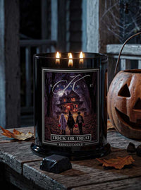A black glass jar candle with three lit wicks sits on a rustic wooden table, surrounded by Halloween props including a carved pumpkin and candy. The front label reads 'Kringle' and 'Trick or Treat' with a dark haunted-house illustration.