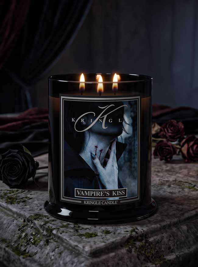 A black glass jar Halloween candle with three lit flames, labeled 'Vampire's Kiss' by Kringle Candle, set on a stone surface with dark roses and draped fabric in the background.