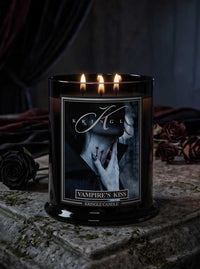 A black glass jar Halloween candle with three lit flames, labeled 'Vampire's Kiss' by Kringle Candle, set on a stone surface with dark roses and draped fabric in the background.