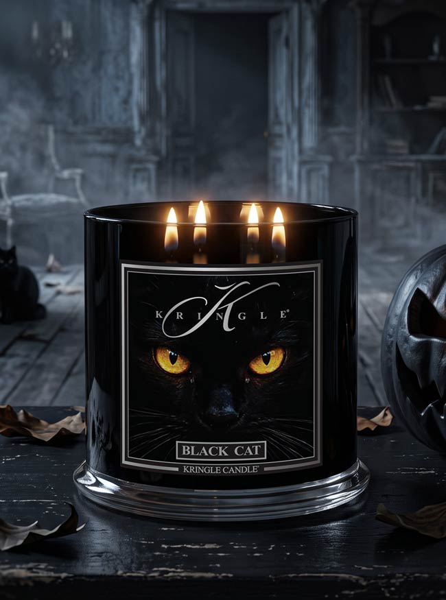 A black glass Kringle candle labeled 'Black Cat' with three lit candles, shown on a dark tabletop in a moody, gothic room setting with a carved pumpkin partially visible at the side.