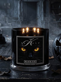 A black glass Kringle candle labeled 'Black Cat' with three lit candles, shown on a dark tabletop in a moody, gothic room setting with a carved pumpkin partially visible at the side.