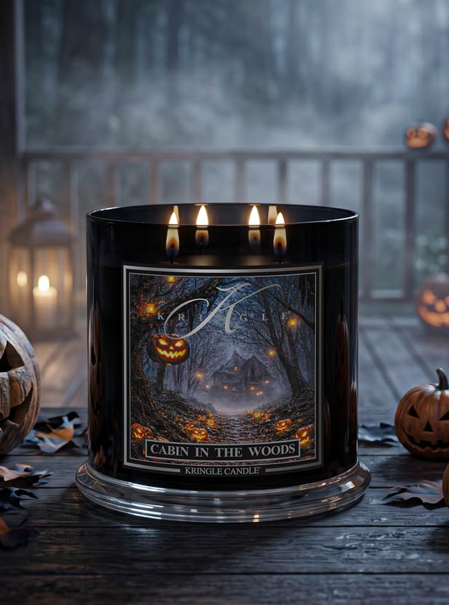 A black Kringle Candle jar with three lit wicks, featuring a Halloween-themed label that reads 'Cabin in the Woods.' The candle sits on a dark wooden surface with pumpkins and lanterns blurred in the background.