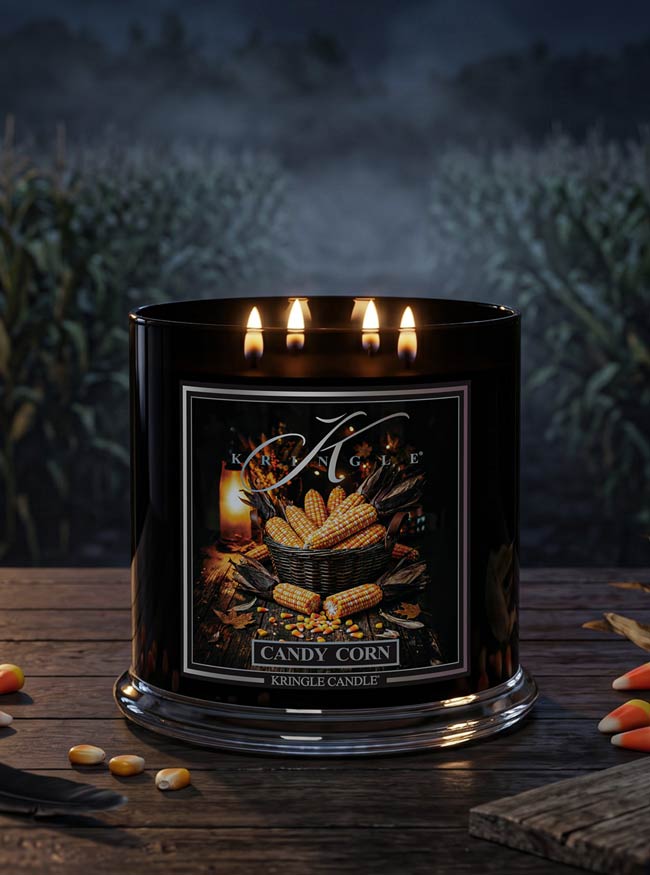 A black three-wick scented candle labeled 'Candy Corn' by Kringle Candle, lit and set on a rustic wooden table with corn kernels and cornstalks scattered around, against a dark, misty field background.