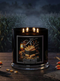 A black three-wick scented candle labeled 'Candy Corn' by Kringle Candle, lit and set on a rustic wooden table with corn kernels and cornstalks scattered around, against a dark, misty field background.