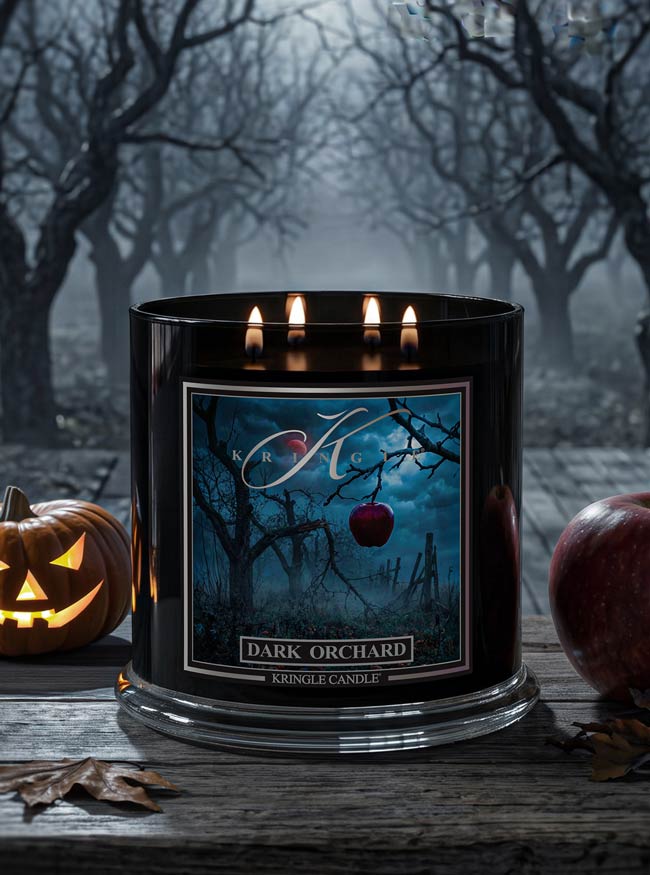 A black Kringle Candle jar labeled 'Dark Orchard' with three lit wicks, displayed on a rustic wooden surface with Halloween props including a jack-o'-lantern, an apple, and a blurred forest scene in the background.