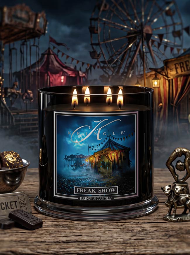 A black Kingle Candle jar labeled 'Freak Show' with four lit wicks, placed on a rustic wooden surface in a carnival-themed scene with a ferris wheel and tents in the background.
