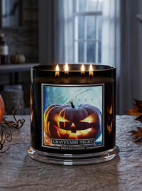 A black three-wick candle labeled 'Graveyard Night' by Kingle Candle, shown lit on a stone surface with autumn leaves and a pumpkin nearby in a softly blurred indoor setting.