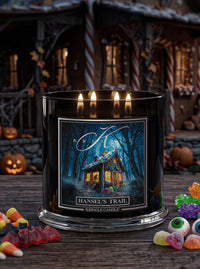 A black glass jar candle with three lit wicks sits on a wooden table in a Halloween-themed setting. The front label reads 'KRingle Candle' and 'Hansel's Trail,' showing an illustration of a haunted house in a dark forest.